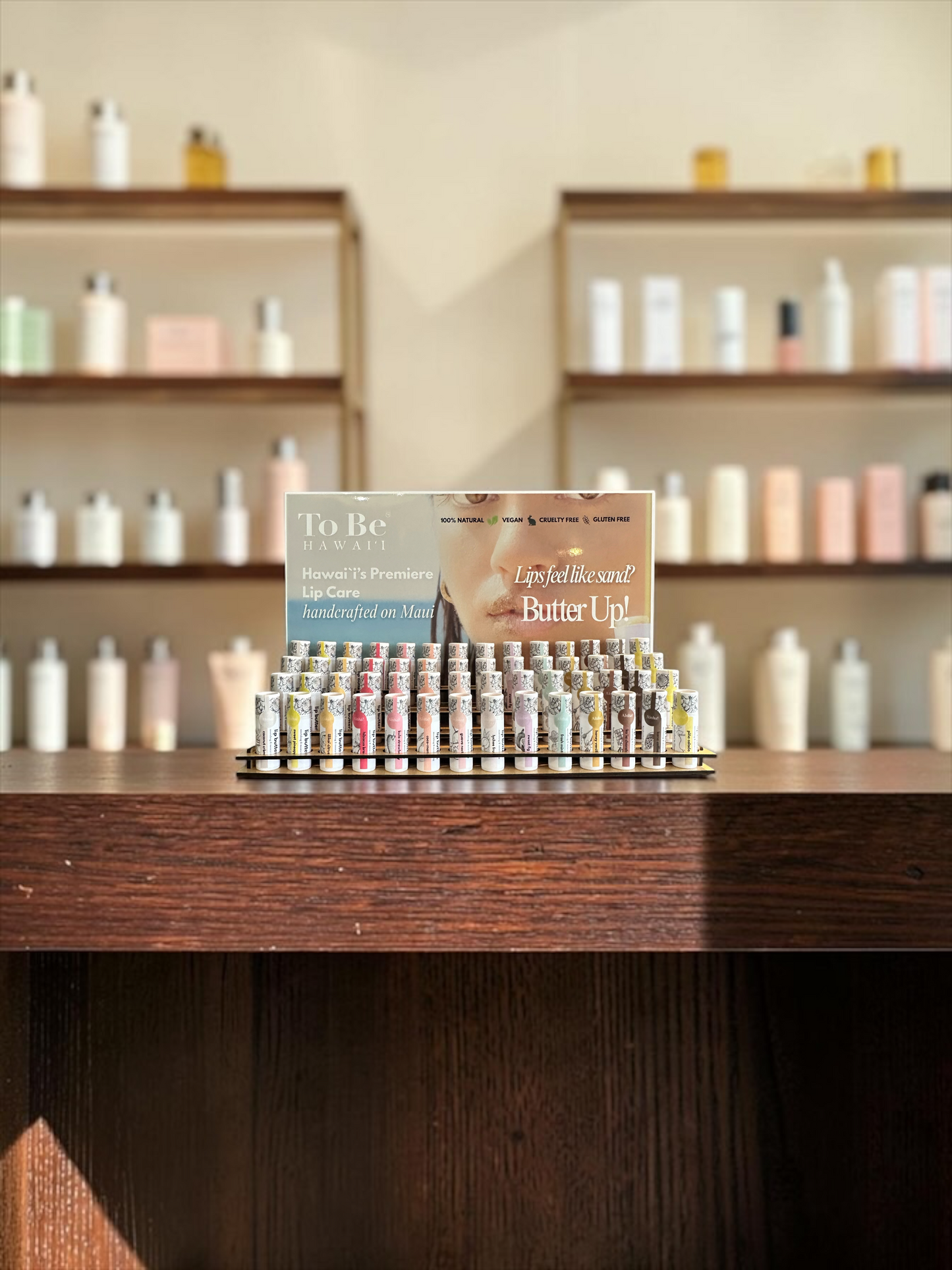 A display of small cosmetic vials arranged in a pyramid on a wooden counter, with a sign reading 'To Be Natural' and 'Lip Tinted Butter Up!' behind them. Shelves with assorted bottles are blurred in the background.