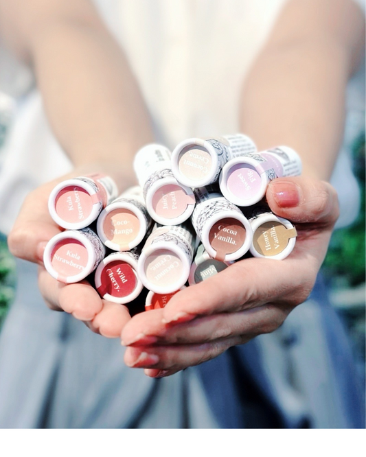 A person holds a cluster of lip gloss tubes in both hands. The tubes feature a white label with black patterned text and display various pastel and neutral shades, including pink, nude, beige, and red tones.