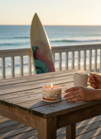 Candle and mug of coffee  on a wooden table with a beach view and a surfboard in the background.