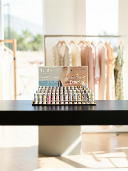 A display of lip butters reading 'To Be Hawaii' and ' Butter Up!' in a bright, softly blurred boutique interior.