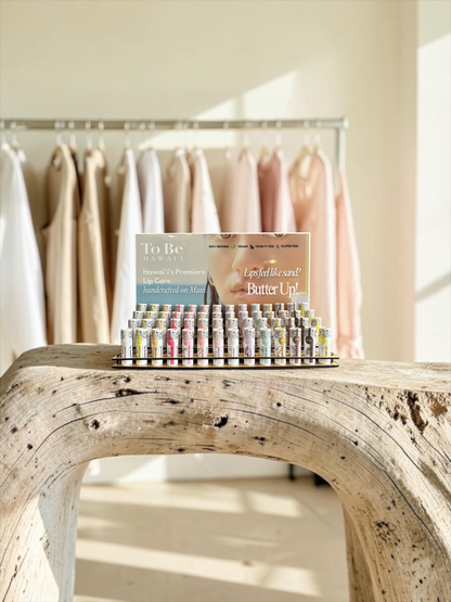 Full lip balm display fill with To Be Hawaii Lip Butters on a driftwood wood retail shelf with clothing rack behind it.