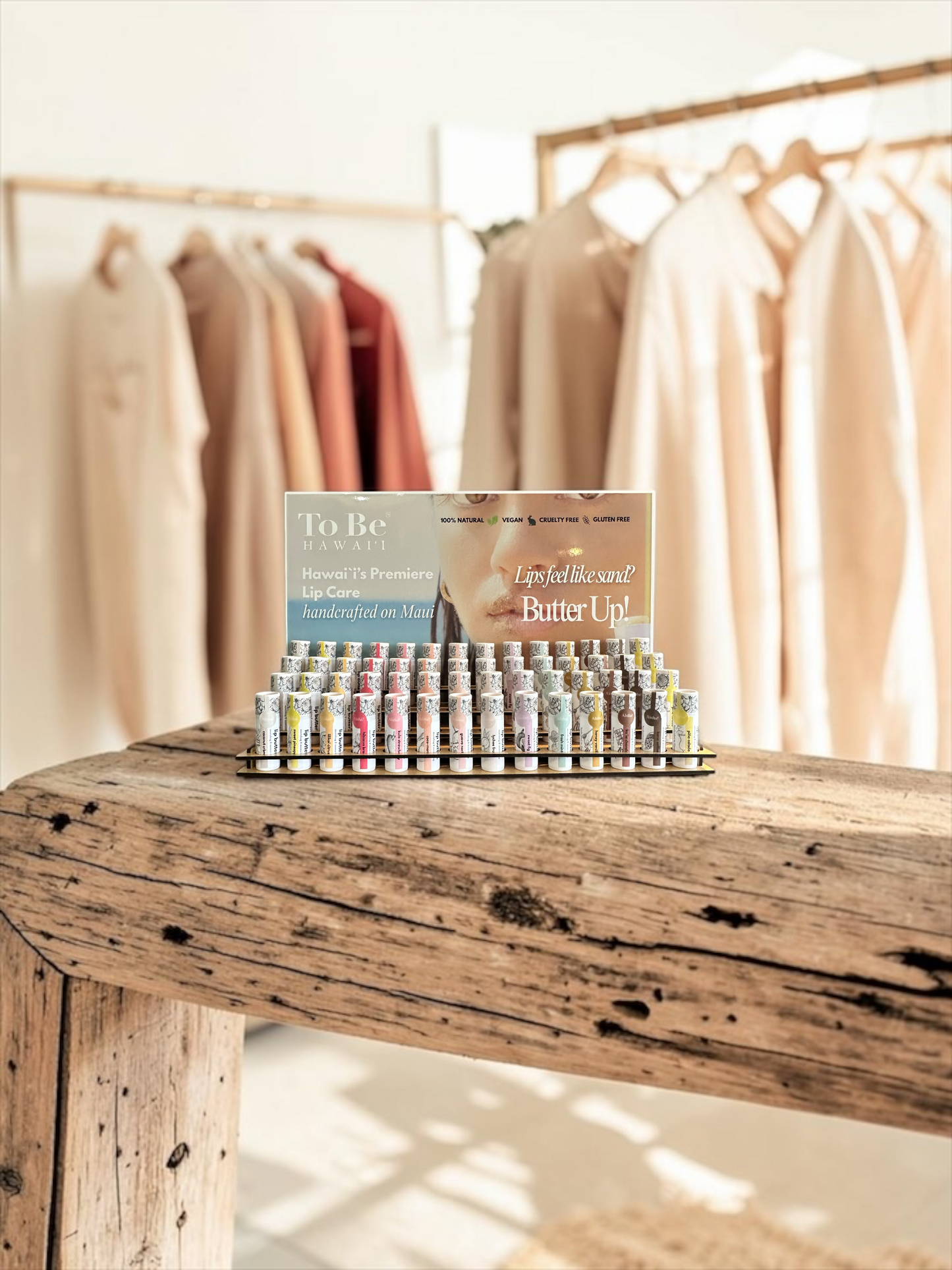 A small product display of lip balm tubes and a branded card reading 'To Be Hawaii' and 'Lip Balm with Butter Up!' sits on a rustic wooden table in a softly lit boutique interior with hanging neutral-toned garments in the background.