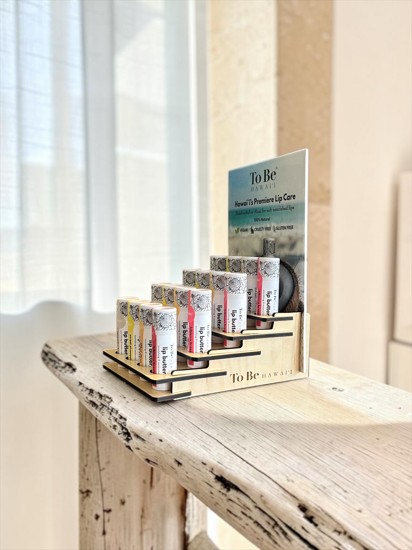 A wooden display stand holding several lip balm tubes from To Be Hawaii, with a larger product card behind them, placed on a rustic wooden table in a softly lit indoor setting.