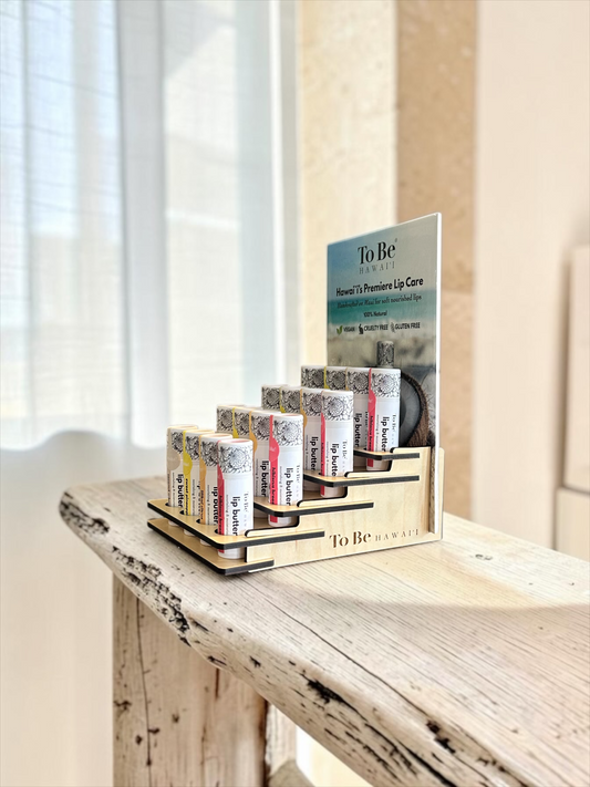 A wooden display stand holding several lip balm tubes from To Be Hawaii, with a larger product card behind them, placed on a rustic wooden table in a softly lit indoor setting.