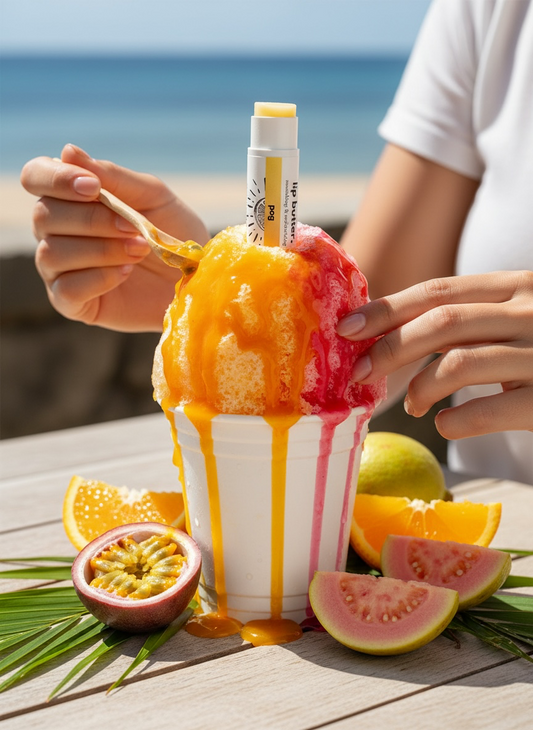 Person holding a colorful shaved ice with a beach in the background. The POG flavor lip butter is sticking out of the shaved ice.