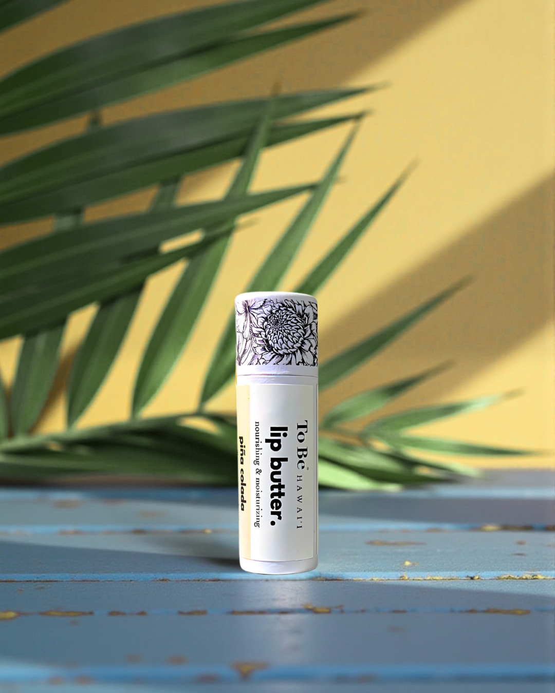 "to Be" branded pina colada Lip butter container on a wooden surface with a yellow wall and green leaves in the background