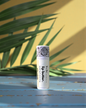 "to Be" branded pina colada Lip butter container on a wooden surface with a yellow wall and green leaves in the background