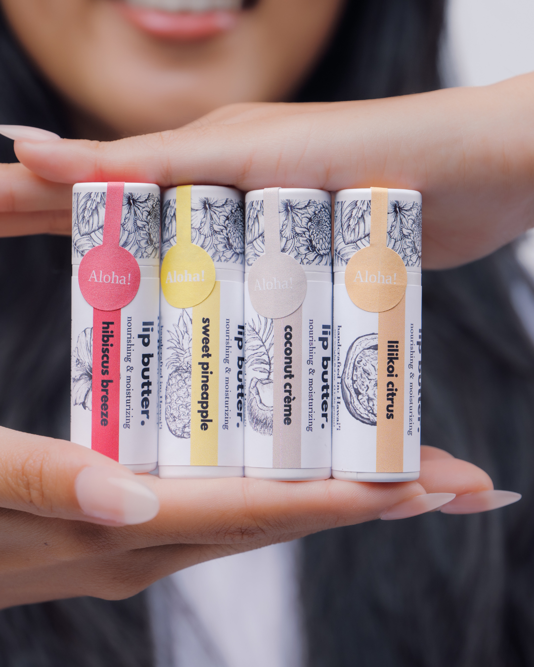 A close-up of a person holding five lip balm tubes in their hands. The lip balms are arranged horizontally, showing different flavor labels: Hibiscus breeze (pink), sweet pineapple (yellow), coconut cream (tan), and lilikoi citrus (peach).