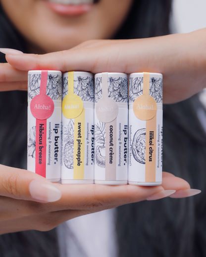 A close-up of a person holding five lip balm tubes in their hands. The lip balms are arranged horizontally, showing different flavor labels: Hibiscus breeze (pink), sweet pineapple (yellow), coconut cream (tan), and lilikoi citrus (peach).