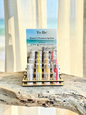 A set of lip balm tubes from To Be Beautiful Hawaii's Premier Lip Care displayed on a wooden stand on a rustic wood slab, with a beach-themed background and sheer curtains.