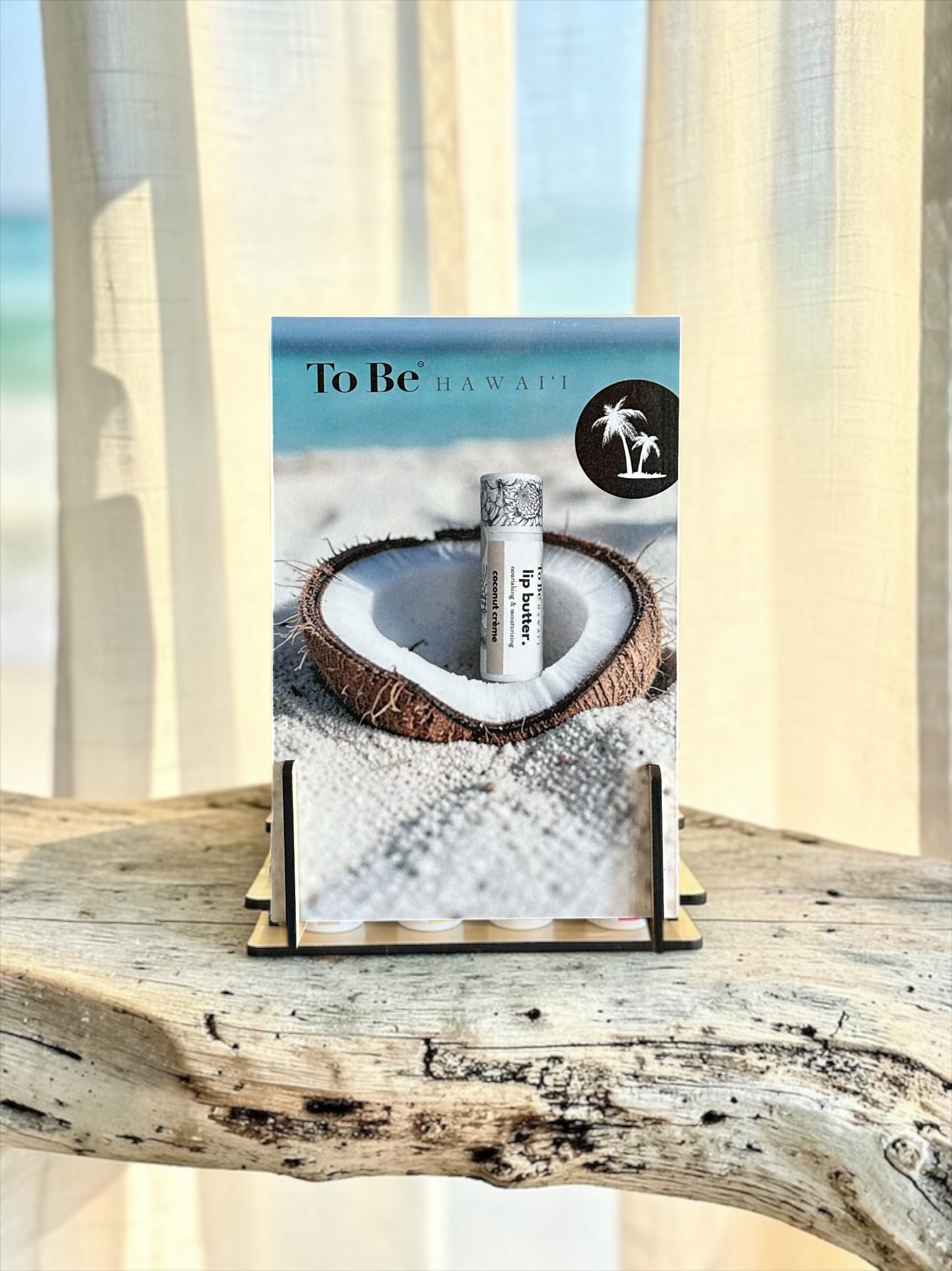 A lip balm tube labeled 'To Be HAWAII'  displayed on a small wooden stand. The packaging features a tropical beach scene with a coconut shell on white sand, set against a background of draped cream curtains and a rustic wooden surface.